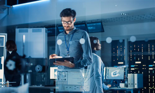 Chief Engineer Stands in the Middle of the Lab and Uses Tablet Computer. Team of Industrial Engineers, Developers Work on Engine Design Use Digital Whiteboard and Computers