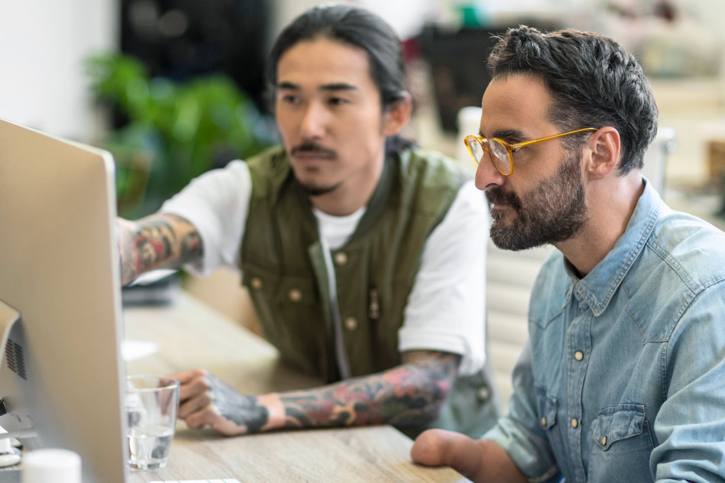 Two employees of a digital business work on their WordPress site wearing casual clothing, one has glasses with a beard, another has tattooed arms with a mustache