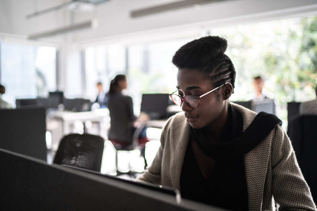 A woman in stylish, thin-rimmed glasses and short hair at her office, she compares Magento hosting plans while her coworkers work in the background, she is wearing a crossbody bag