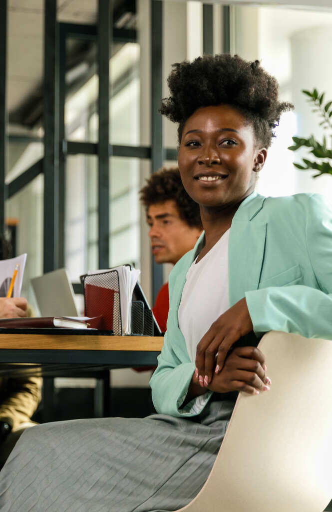 A woman in a blazer and short hair sits sideways on a chair and smiles as her coworkers chat about choosing WordPress VPS