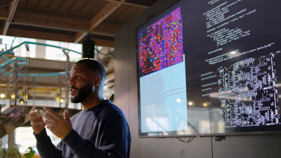 an instructor with a light beard giving a lecture at an educational institution in front of a large monitor, which shows code and an engineering blueprint