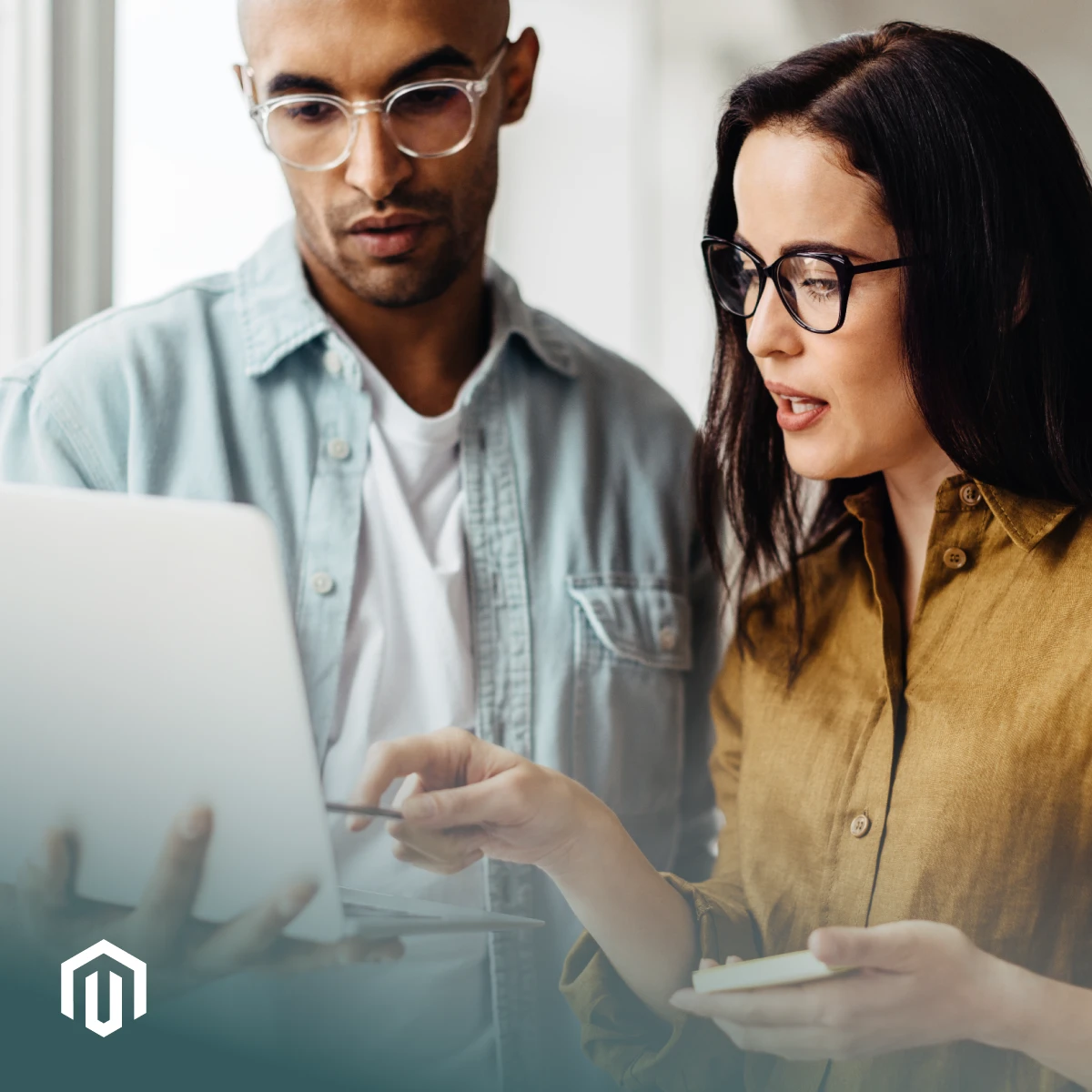 Man and woman wearing glasses and button down shirts stand looking at a laptop together to assess their Magento fulfillment needs