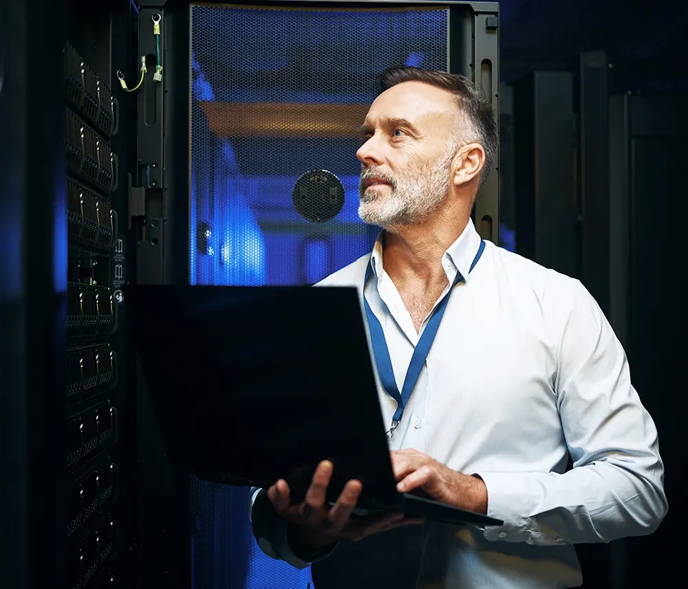 A hosting technician wearing a badge holds his computer as he monitors server performance in a data center hall
