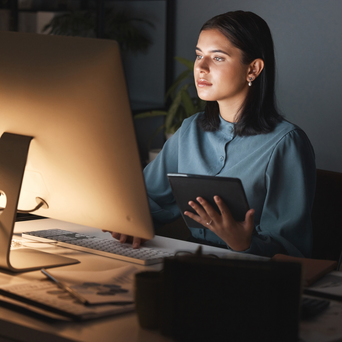 A woman with shoulder-length hair gazes at her monitor while building a WordPress website and selecting plugins. She holds a tablet device in one hand while using her computer mouse.
