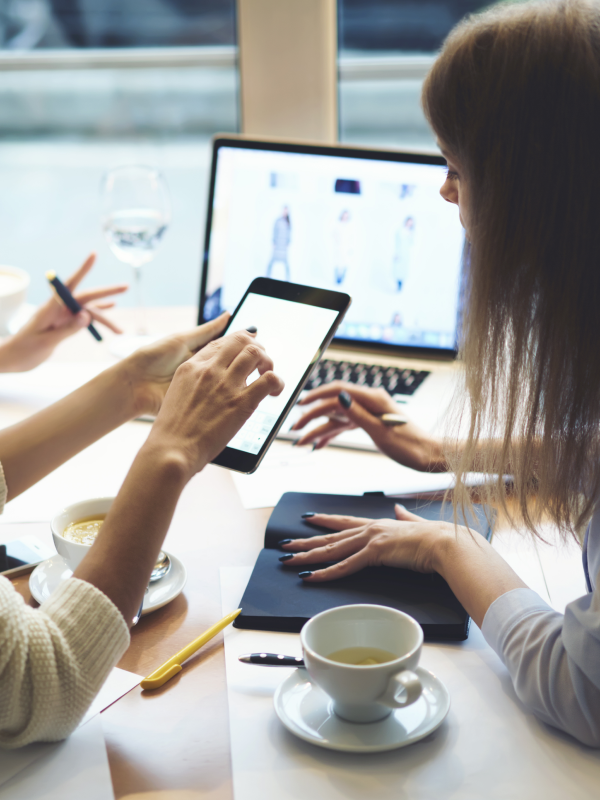 A woman and her friends at a coffee shop gaze at a phone used to shop using AI. A laptop is open on the table with a traditional ecommerce store selection.