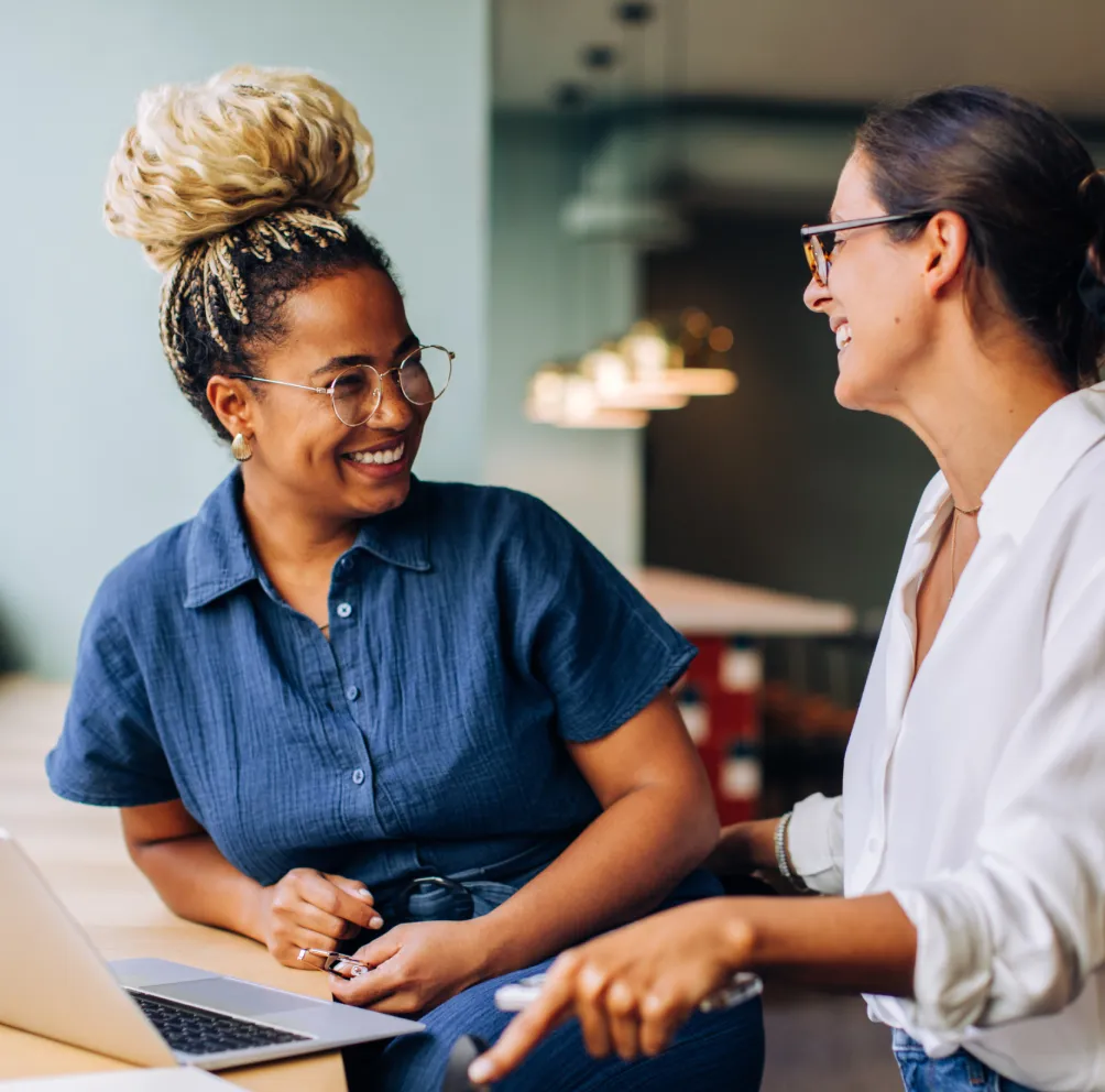Two women smile as they collaborate, one woman is leaning on a countertop, a laptop is open on the counter.